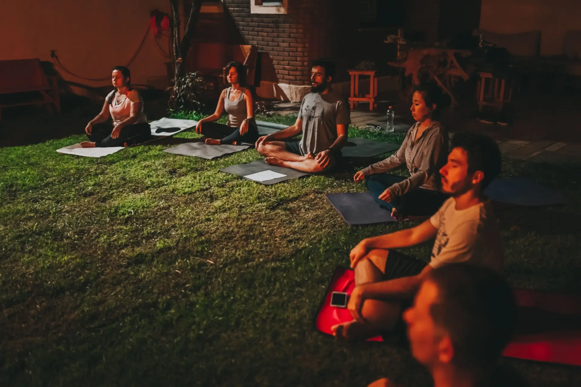 A diverse group meditating outdoors on yoga mats during evening time.