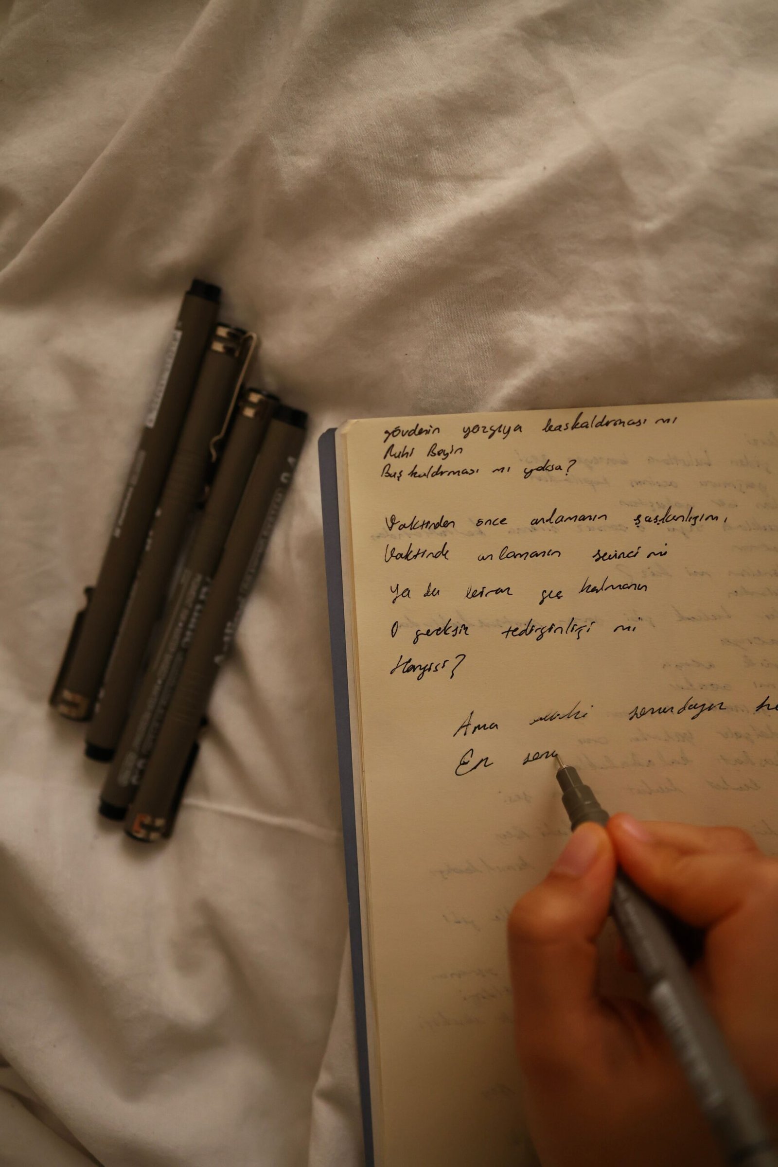 A hand writes in a notebook with pens nearby on a cozy bed setting.